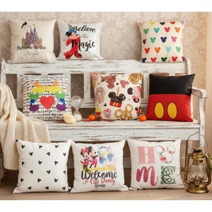 Könnte beinhalten: Eine Sammlung dekorativer Zierkissen mit verschiedenen Designs, darunter Mickey Mouse, Schloss- und Herzmuster. Einige Kissen zeigen Texte wie "Believe in the Magic" und "Welcome to Our Disney Home". Die Kissen sind auf einer weißen Bank angeordnet.