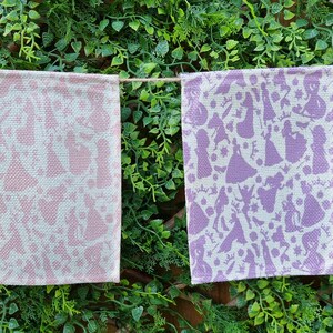 May include: A string of four fabric pennants with a white background and a repeating pattern of purple, pink, and red silhouettes of princesses. The pennants are hanging from a string against a backdrop of green foliage.
