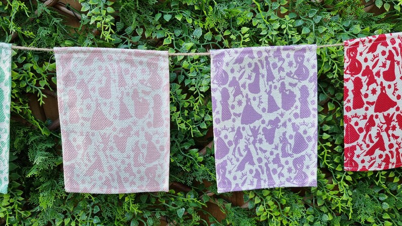 May include: A string of four fabric pennants with a white background and a repeating pattern of purple, pink, and red silhouettes of princesses. The pennants are hanging from a string against a backdrop of green foliage.