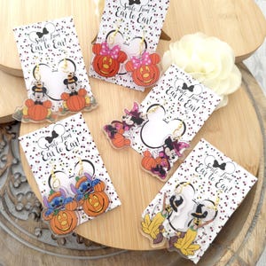 May include: Five pairs of acrylic dangle earrings featuring Disney characters in Halloween costumes, including Mickey and Minnie Mouse, Stitch, and Pluto with pumpkins. Each pair is attached to a white display card with the text Smile from Ear to Ear.