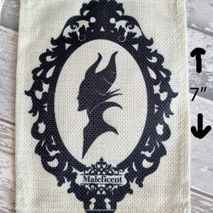 May include: A burlap banner with a black silhouette of Maleficent in a decorative frame. The banner is 5 inches wide and 7 inches tall. The text "Maleficent" is printed below the frame.