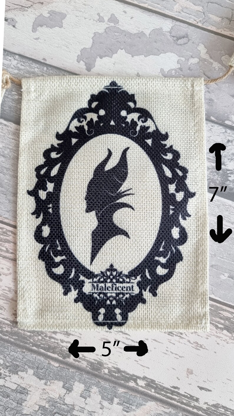 May include: A burlap banner with a black silhouette of Maleficent in a decorative frame. The banner is 5 inches wide and 7 inches tall. The text "Maleficent" is printed below the frame.
