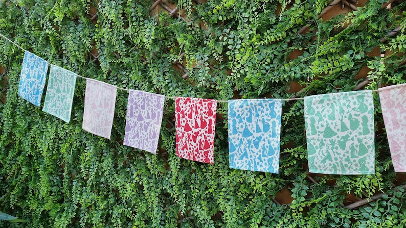 May include: A string of colorful fabric flags with a white background and a repeating pattern of white silhouettes of people dancing. The flags are strung on a white cord and hanging against a green leafy background.