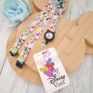 Może przedstawiać: Smycz z kolorowym wzorem z postaciami z kreskówek i pasującym uchwytem na identyfikator. Uchwyt na identyfikator zawiera tekst "My 1st Disney Visit" z uszami Myszki Miki i fajerwerkami. Smycz znajduje się na drewnianej desce w kształcie Myszki Miki.