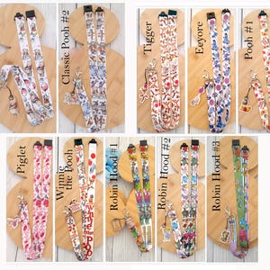 May include: A collection of ten fabric lanyards featuring various character prints including Winnie the Pooh, Tigger, Eeyore, Piglet, and Robin Hood. Each lanyard includes a metal lobster clasp and a matching character charm attached to the end.