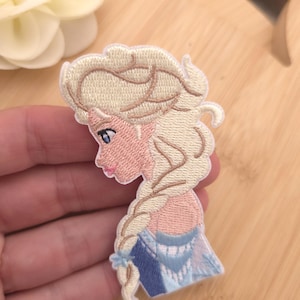 Könnte beinhalten: Ein gestickter Aufnäher mit dem Seitenprofil einer Figur mit langem, geflochtenem blondem Haar, heller Haut und blauen Augen. Die Figur trägt ein blau-weißes Kleidungsstück mit komplexen Stickdetails in den Farben Blond, Blau und Rosa.