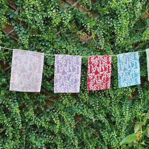 May include: A string of colorful fabric flags with a white pattern of stylized figures hanging against a green leafy background.
