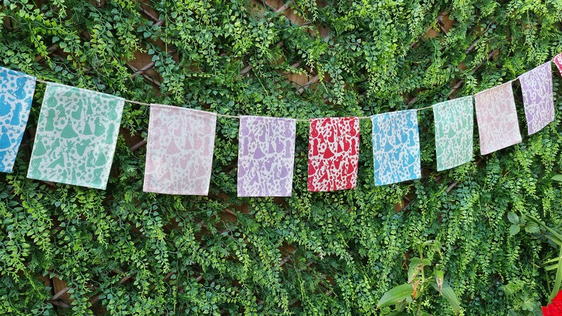 May include: A string of colorful fabric flags with a white pattern of stylized figures hanging against a green leafy background.