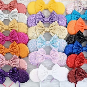 May include: A set of 16 pairs of sparkly mouse ears with bows in various colours, including red, orange, yellow, green, blue, purple, pink, and white. The ears are made of sequins and are attached to a headband.