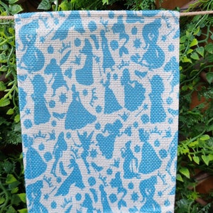 May include: A white burlap flag with a blue pattern of various silhouettes. The silhouettes are of people in dresses and other figures.