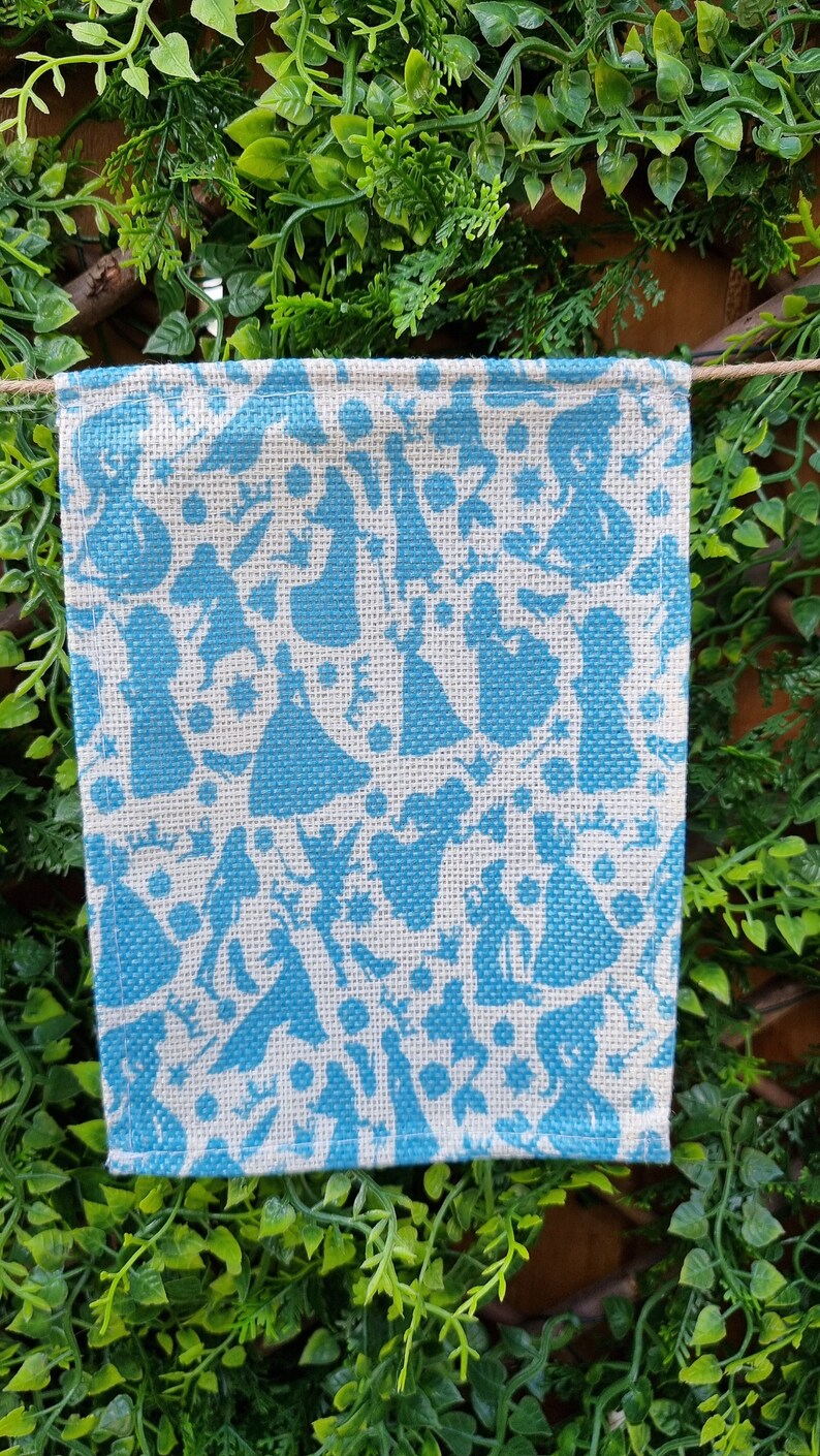May include: A white burlap flag with a blue pattern of various silhouettes. The silhouettes are of people in dresses and other figures.