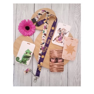 May include: A purple lanyard with a white and gold pattern featuring Disney characters. The lanyard has a gold charm with a sun design. There are four white card holders with different Disney character images. One card holder has a green chameleon, another has a sun, another has Rapunzel and Flynn Rider, and the last one has Rapunzel's hair.
