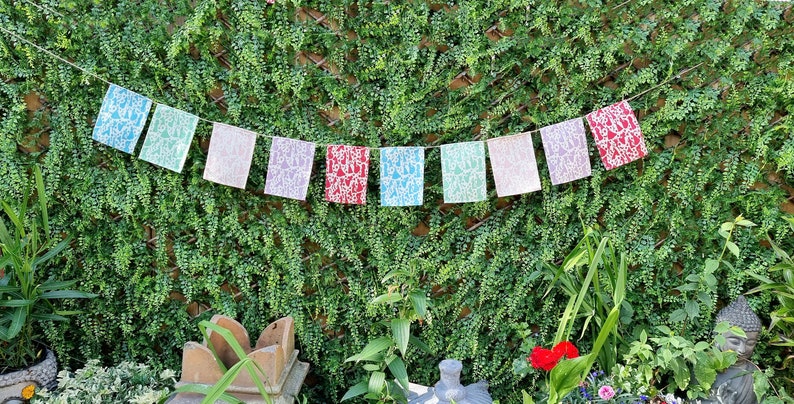 May include: A string of colorful prayer flags with white designs hanging against a green leafy wall. The flags are in shades of blue, green, pink, red, and white.
