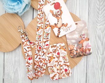 May include: Two white and orange lanyard keychains with a cartoon image of Tigger from Winnie the Pooh. The lanyards have a black clip and a white and orange ID card holder with a cartoon image of Tigger. The ID card holder has the word "Tigger" printed on it.