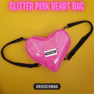 May include: Pink heart-shaped crossbody bag with glitter finish and black straps. The bag has a white label with the text "Grouchbag" on it.