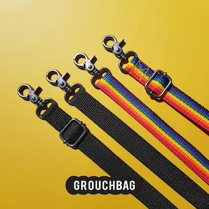 May include: Four different camera straps, two are black with silver clasps and two are rainbow striped with silver clasps. The straps are laid out on a yellow surface. The text "Grouchbag" is printed on the surface.