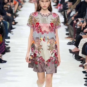 May include: A woman wearing a short-sleeved, floral patterned dress with a sheer overlay. The dress has a beaded neckline and a layered skirt. She is wearing pink sandals with a bow detail.