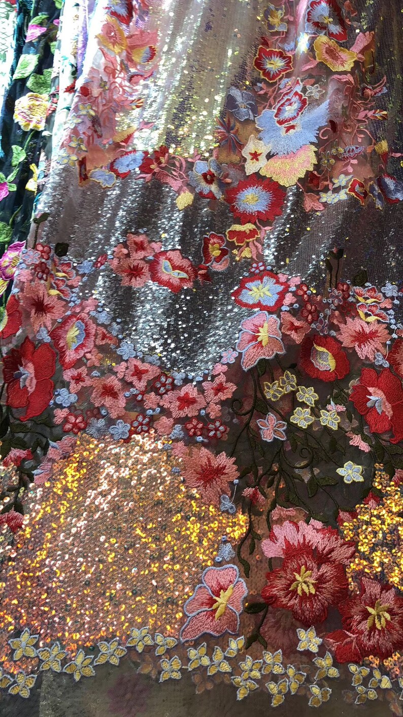 May include: A close-up of a fabric with a shimmering gold and silver sequin background and a floral embroidery pattern in shades of pink, red, blue, and yellow.