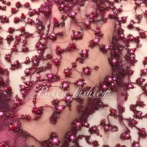 Op de afbeelding: Close-up van bordeauxrode stof met bloemige kralen- en paillettenversieringen. Het ontwerp heeft clusters van roze en rode kralen, met de tekst "Bella fashion" in een cursief lettertype.