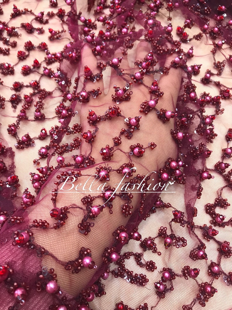 Op de afbeelding: Close-up van bordeauxrode stof met bloemige kralen- en paillettenversieringen. Het ontwerp heeft clusters van roze en rode kralen, met de tekst "Bella fashion" in een cursief lettertype.