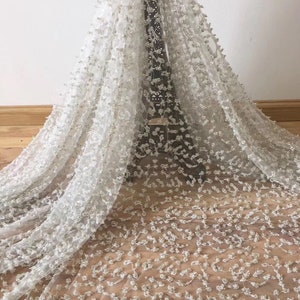 Op de afbeelding: Witte kanten stof met een bloemmotief en verspreide pailletten. Het delicate materiaal is over een houten oppervlak gedrapeerd, wat de textuur en het ontwerp laat zien. Ideaal voor bruidskleding of speciale gelegenheidskleding.