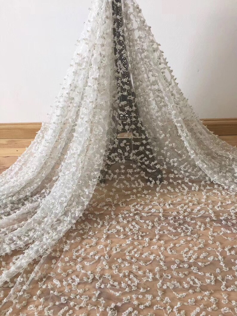 Op de afbeelding: Witte kanten stof met een bloemmotief en verspreide pailletten. Het delicate materiaal is over een houten oppervlak gedrapeerd, wat de textuur en het ontwerp laat zien. Ideaal voor bruidskleding of speciale gelegenheidskleding.