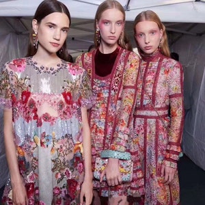 May include: Three women wearing colorful floral patterned dresses. The dresses are made of a sheer fabric with embroidery and sequins. The women are standing in a white tent.