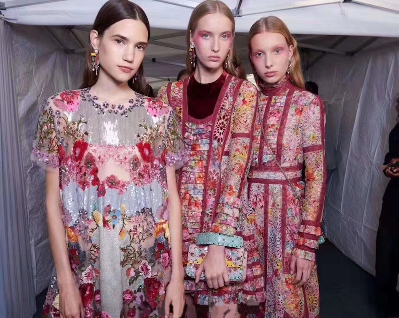 May include: Three women wearing colorful floral patterned dresses. The dresses are made of a sheer fabric with embroidery and sequins. The women are standing in a white tent.
