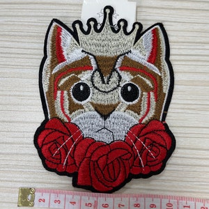 May include: Embroidered iron-on patch of a cat wearing a crown and surrounded by red roses. The patch is approximately 5 inches wide.