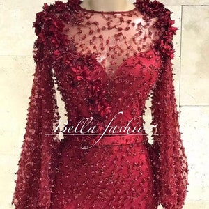 Op de afbeelding: Een formele, bordeauxrode jurk met lange mouwen. De jurk heeft ingewikkelde bloemversieringen op de schouders en het lijfje, met een doorschijnende halslijn en een getailleerd silhouet. De tekst "Bella fashion" is zichtbaar.