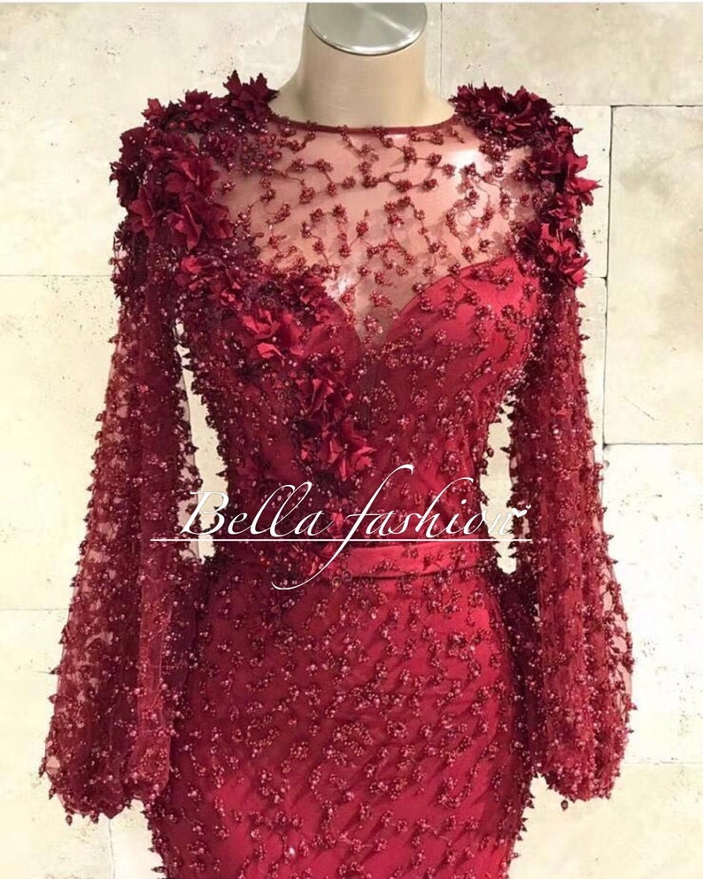 Op de afbeelding: Een formele, bordeauxrode jurk met lange mouwen. De jurk heeft ingewikkelde bloemversieringen op de schouders en het lijfje, met een doorschijnende halslijn en een getailleerd silhouet. De tekst "Bella fashion" is zichtbaar.