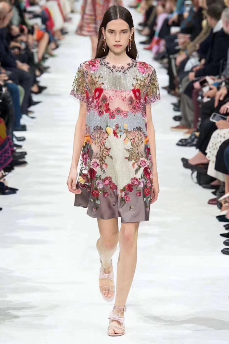 May include: A woman wearing a short-sleeved, floral patterned dress with a sheer overlay. The dress has a beaded neckline and a layered skirt. She is wearing pink sandals with a bow detail.