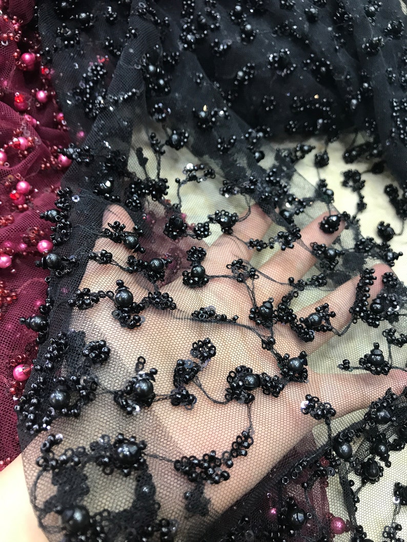 Op de afbeelding: Close-up van zwarte geborduurde kantstof. Het delicate gaas is versierd met clusters van zwarte kralen en pailletten, waardoor een textuurpatroon ontstaat. Een gedeelte van bordeauxrode stof met roze kralen is zichtbaar op de achtergrond.