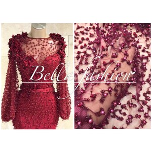 Op de afbeelding: Een bordeauxrode avondjurk met lange mouwen en een doorschijnende halslijn, versierd met ingewikkeld kralenwerk en bloemmotieven. De afbeelding toont ook een close-up van de kralenstof. De tekst "Bella fashion" staat over de afbeelding.