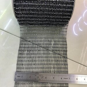 Puede incluir: Un rollo de ribete negro de strass con strass transparentes. El ribete se mide con una regla que muestra 7 pulgadas.