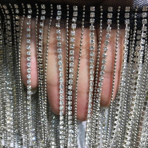 May include: A close-up of a black mesh trim with silver chains and clear rhinestones. The trim is made of a fine mesh fabric with a fringe of silver chains and clear rhinestones. The rhinestones are square-shaped and are evenly spaced along the chains.