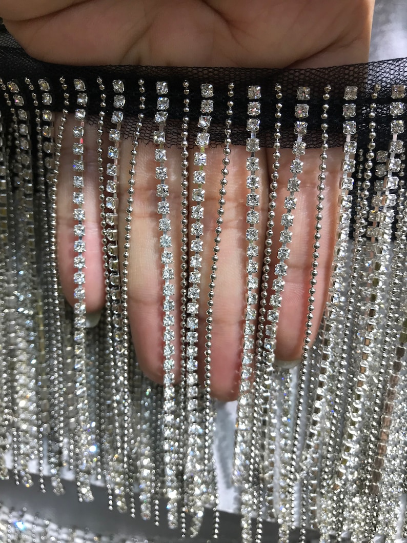 May include: A close-up of a black mesh trim with silver chains and clear rhinestones. The trim is made of a fine mesh fabric with a fringe of silver chains and clear rhinestones. The rhinestones are square-shaped and are evenly spaced along the chains.