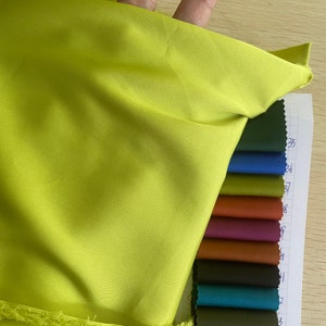May include: A bright yellow fabric swatch with a frayed edge. The swatch is held in a hand and is next to a colour chart of various shades of green, blue, brown, and red.