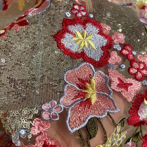May include: Close-up of a sheer fabric with embroidered floral designs in shades of pink, red, yellow, and blue. The fabric is covered in silver sequins.