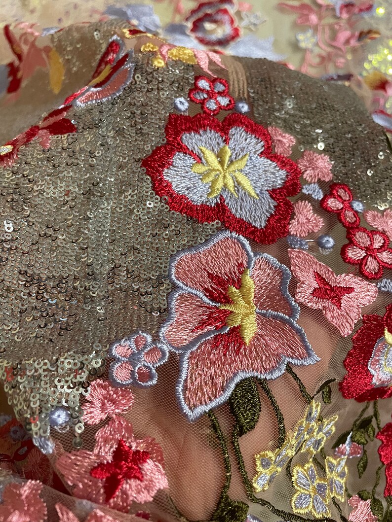 May include: Close-up of a sheer fabric with embroidered floral designs in shades of pink, red, yellow, and blue. The fabric is covered in silver sequins.