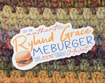 Könnte beinhalten: Ein gestanzter Aufkleber mit einem Cartoon-Hamburger und dem Text: TRY AN authentic Ryland Grace MEBURGER MADE WITH 100% ETHICALLY SOURCED (Me)at. Der Aufkleber hat einen weißen Rand und liegt auf einem mehrfarbigen, strukturierten Häkelhintergrund.