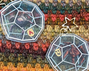 May include: Two clear acrylic keychains with a geometric pentagonal frame design. One features a dark figure with a red heart icon, the other a red figure with a yellow musical note icon. Both have gold-toned star-shaped clasps at the top.