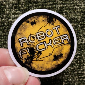 以下が含まれることがあります： 黒い縁取りと白いブロック体の「ROBOT FUCKER」の文字が入った丸いステッカー。背景は黄色で、黒い飛沫と回路のデザインが施されています。