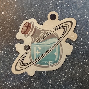 May include: A sticker featuring a potion bottle with a cork stopper, surrounded by a planet's rings. The bottle contains a teal liquid with cross-shaped sparkles. The sticker has a white border and is set against a starry background.