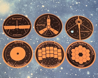 Puede incluir: Seis pegatinas circulares con dibujos de líneas naranjas de misiones espaciales sobre un fondo negro. Las pegatinas están etiquetadas como Europa Clipper, Juno, Voyager, Hubble Space Telescope, Nancy Grace Roman Space Telescope y James Webb Space Telescope.