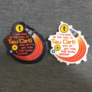 Puede incluir: Dos pegatinas con tem&aacute;tica espacial. Cada pegatina presenta un cohete con llamas naranjas y rojas, el texto "I traveled all the way to Tau Ceti and all I got was this lousy sticker" y un c&iacute;rculo amarillo con un signo m&aacute;s.