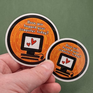 以下が含まれることがあります： 「Have you hugged your computer today?」の白文字が入った丸いステッカー2枚。ステッカーには、2つの赤いハートが付いたコンピューターのグラフィックが描かれています。背景はオレンジ色で、デジタル回路のデザインです。