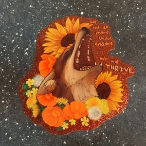 May include: A sticker featuring a howling wolf with sunflowers and orange flowers. The sticker has the text "We will do more than endure" and "We will thrive" in white font. The sticker has a glittery, reddish-brown border.