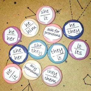May include: A set of 10 round buttons with different pronoun options, including "he/him", "she/her", "they/them", "she/they", "he/they", "it/its", "any pronouns", and "ask for pronouns". The buttons are in various colors, including pink, blue, and beige.