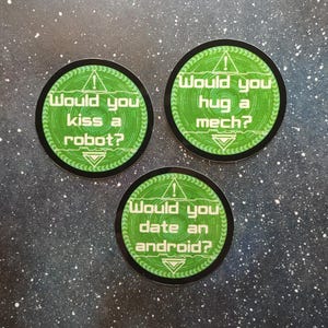 以下が含まれることがあります： 緑と黒のデザインの丸いステッカーが3枚。各ステッカーには、白い文字で異なる質問が書かれています。「ロボットにキスしますか？」「メカを抱きしめますか？」、そして「アンドロイドとデートしますか？」ステッカーは、暗く斑点のある背景にあります。